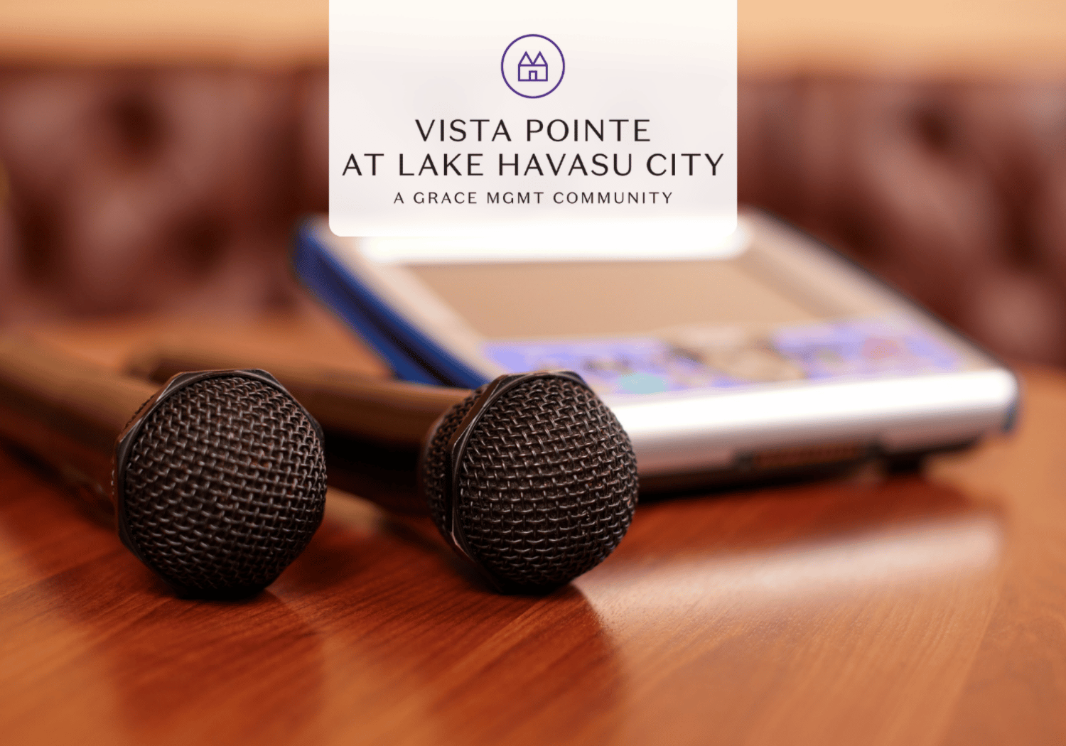 Two black microphones sit on a polished wooden table with a blurred device in the background and a white logo card reading 'Vista Pointe at Lake Havasu City' centered above.